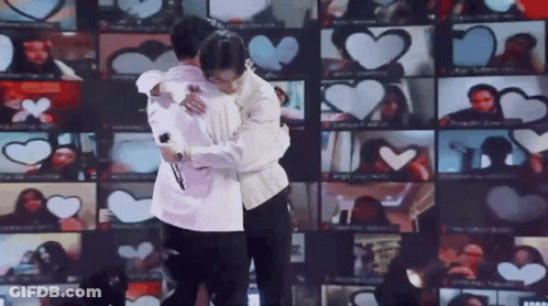 Two Men Are Hugging Each Other In Front Of A Wall Of Hearts . GIF