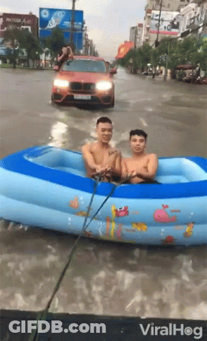 Two Men Are In An Inflatable Pool In A Flooded Street With A Car Behind Them GIF
