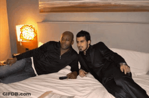 Two Men Are Laying On A Bed And One Has A Remote Control In His Hand GIF
