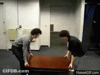 Two Men Are Lifting A Wooden Table In A Room . GIF