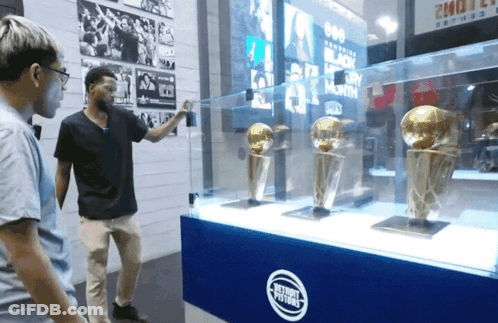 Two Men Are Looking At Trophies In A Glass Case That Says Detroit Pistons GIF