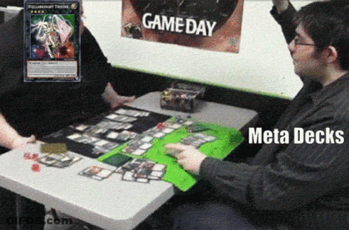 Two Men Are Playing A Game Of Yu Gi Oh With A Game Day Poster Behind Them GIF