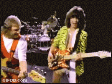 Two Men Are Playing Guitars On A Stage With A Drum Set In The Background GIF