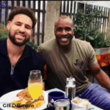 Two Men Are Posing For A Picture While Sitting At A Table With Food And Drinks GIF