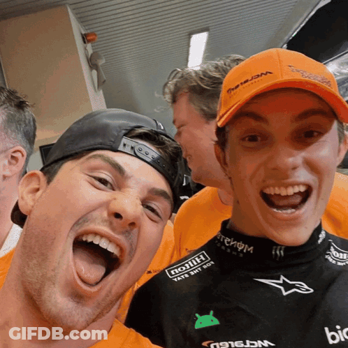 Two Men Are Posing For A Picture With One Wearing An Orange Hat That Says ' Triumph ' On It GIF