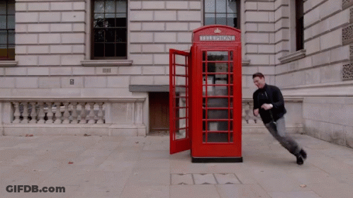 Two Men Are Running In Front Of A Red Telephone Booth With The Door Open GIF