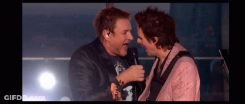 Two Men Are Singing Into Microphones On A Stage While Playing Guitars . GIF