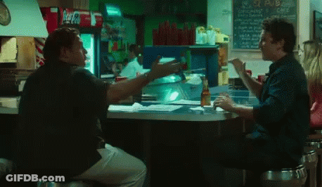 Two Men Are Sitting At A Counter In A Restaurant With A Coca Cola Fridge In The Background GIF
