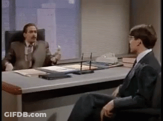 Two Men Are Sitting At A Desk Talking To Each Other . GIF