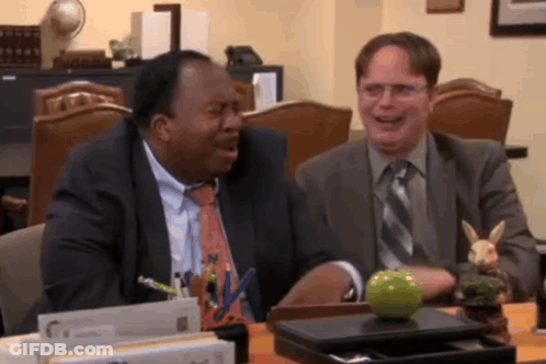 Two Men Are Sitting At A Desk With A Green Apple On It GIF