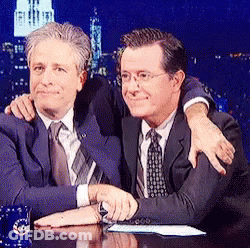 Two Men Are Sitting At A Table And One Has His Arm Around The Other 'S Shoulders GIF