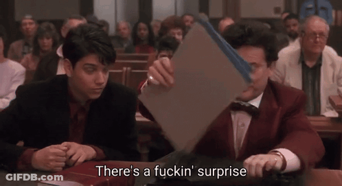 Two Men Are Sitting At A Table And One Of Them Is Holding A Piece Of Paper That Says There 'S A Fuckin Surprise GIF