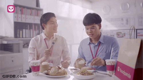 Two Men Are Sitting At A Table Eating Food And Drinking From A Bag That Says Foodpanda GIF