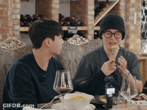 Two Men Are Sitting At A Table Eating Food And Drinking Wine With Bottles Of Wine In The Background GIF
