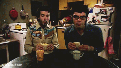 Two Men Are Sitting At A Table Eating Sandwiches In Front Of A Refrigerator With Pictures On It GIF