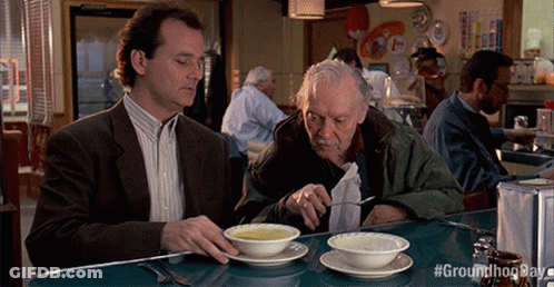 Two Men Are Sitting At A Table Eating Soup With #Groundhogday Written On The Bottom Right GIF