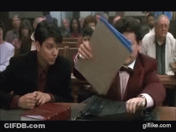 Two Men Are Sitting At A Table In A Courtroom And One Of Them Is Holding A Folder GIF