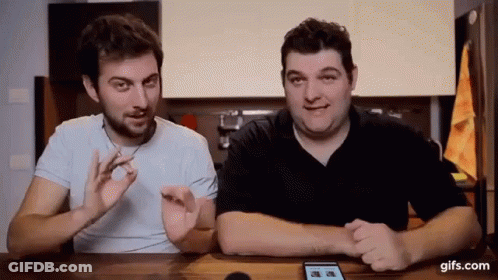 Two Men Are Sitting At A Table Talking To Each Other And Looking At A Phone . GIF