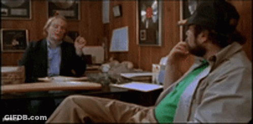 Two Men Are Sitting At A Table Talking To Each Other GIF