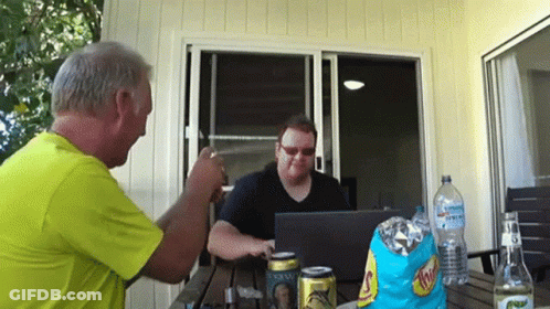 Two Men Are Sitting At A Table With A Bag Of Chips And A Can Of Schweppes On It GIF
