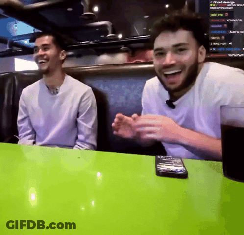 Two Men Are Sitting At A Table With A Green Table Cloth And A Remote Control On It GIF