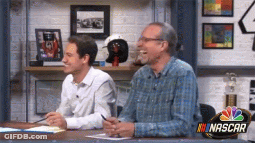 Two Men Are Sitting At A Table With A Nascar Logo On The Wall GIF