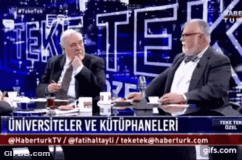 Two Men Are Sitting At A Table With A Sign That Says Tek GIF