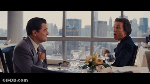 Two Men Are Sitting At A Table With A View Of The City In The Background GIF