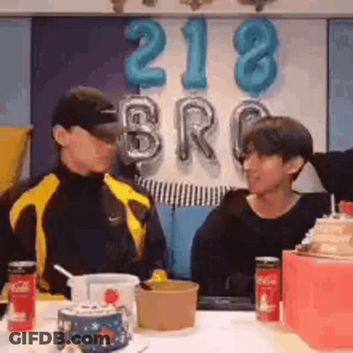 Two Men Are Sitting At A Table With Balloons In The Background And Talking To Each Other . GIF