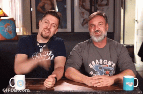 Two Men Are Sitting At A Table With One Wearing A Shirt That Says Dragons On It GIF