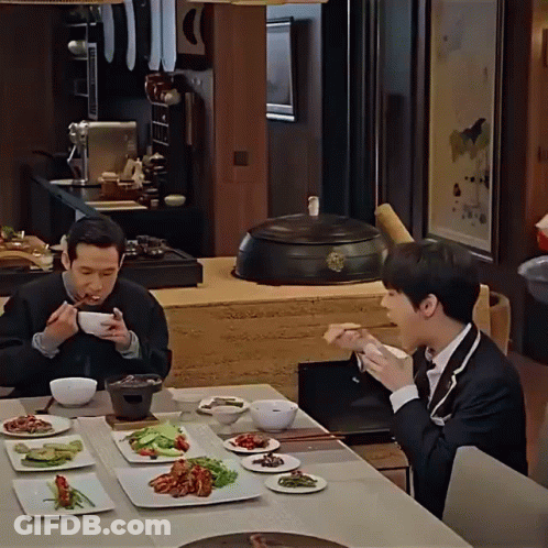 Two Men Are Sitting At A Table With Plates Of Food GIF
