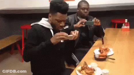 Two Men Are Sitting At A Table With Plates Of Food And One Is Taking A Picture Of The Other . GIF