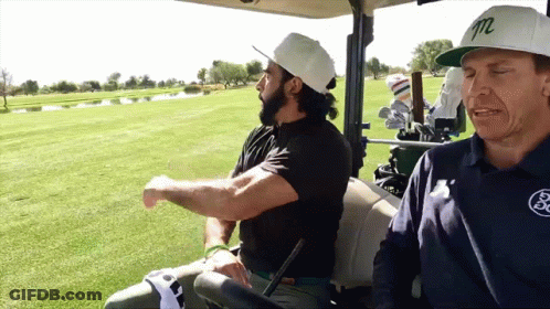 Two Men Are Sitting In A Golf Cart With One Wearing A White Hat With The Letter M On It GIF