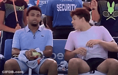 Two Men Are Sitting In A Stadium With A Security Guard In The Background GIF