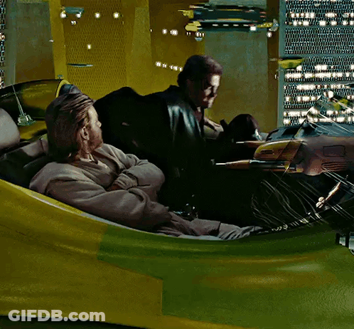 Two Men Are Sitting In A Yellow And Green Vehicle With A City In The Background GIF