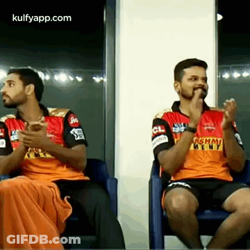 Two Men Are Sitting In Chairs And Clapping Their Hands While Wearing Orange Jerseys GIF