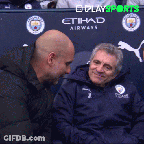 Two Men Are Sitting In Front Of A Manchester City Advertisement GIF