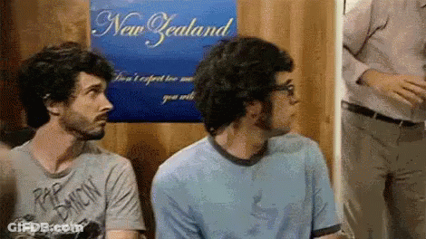 Two Men Are Sitting In Front Of A Poster That Says New Zealand GIF