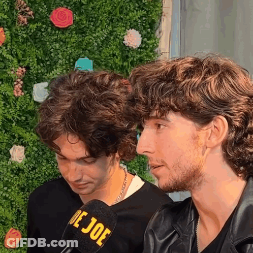 Two Men Are Sitting In Front Of A Wall With Flowers And A Microphone That Says De Joe GIF
