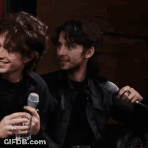 Two Men Are Sitting Next To Each Other Holding Microphones And Drinking . GIF