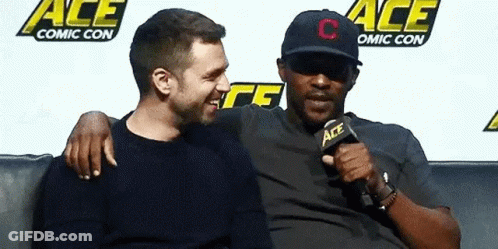 Two Men Are Sitting Next To Each Other On A Couch . One Of The Men Is Wearing A Hat . GIF
