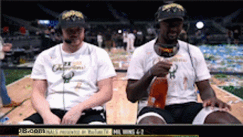 Two Men Are Sitting On A Basketball Court Drinking From A Bottle GIF