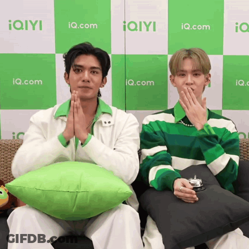 Two Men Are Sitting On A Couch In Front Of A Wall With Iq.com Written On It GIF