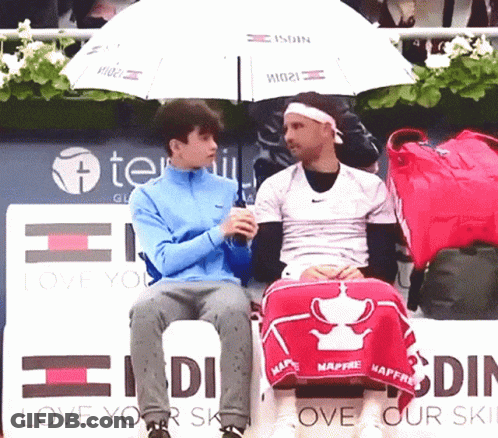 Two Men Are Sitting Under An Umbrella In Front Of A Sign That Says Love Your Skin GIF