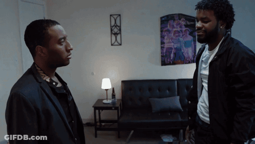 Two Men Are Standing In A Living Room With A Purple Poster On The Wall GIF