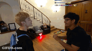 Two Men Are Standing In A Living Room With A Staircase In The Background And One Of Them Has A Backpack That Says Louis GIF