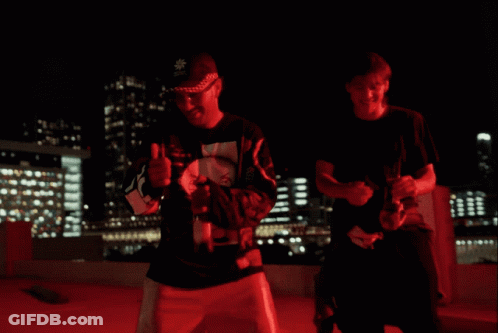 Two Men Are Standing In A Parking Lot At Night With A City In The Background GIF