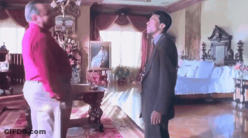 Two Men Are Standing In A Room With A Couch And A Painting On The Wall GIF
