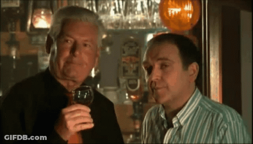 Two Men Are Standing In Front Of A Bar And One Is Holding A Glass Of Wine In His Right Hand GIF