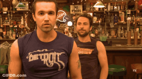 Two Men Are Standing In Front Of A Bar With One Wearing A Shirt That Says Detroit GIF
